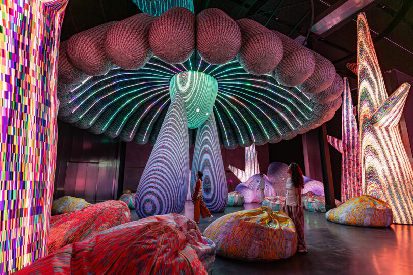 Large illuminated mushroom-shaped sculptures by Nimrod Weis and colorful, textured beanbags fill a dark indoor space, with two people standing and observing the installation.