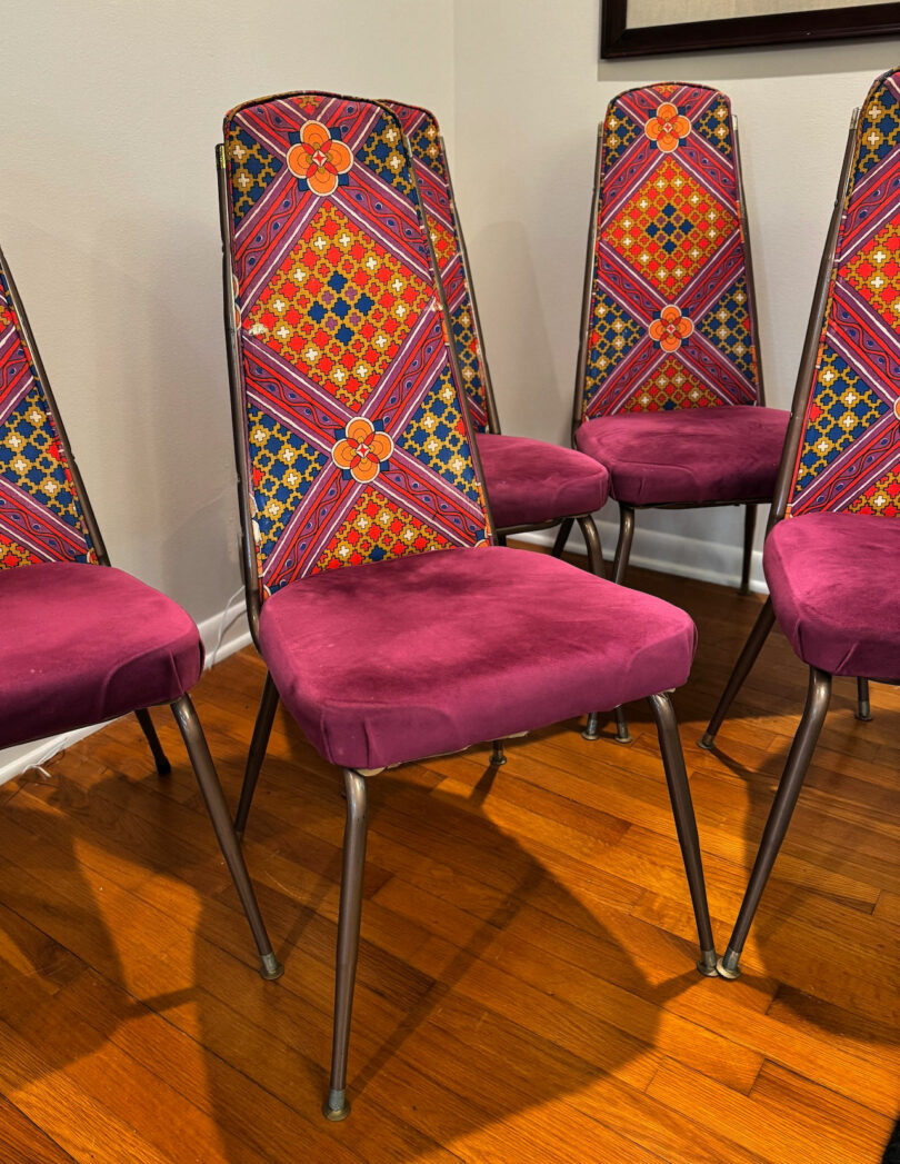 Four metal-framed chairs with purple velvet seats and backs featuring a geometric, multicolored pattern are arranged on a hardwood floor against a white wall—perfect for a stylish Friday 5 gathering.