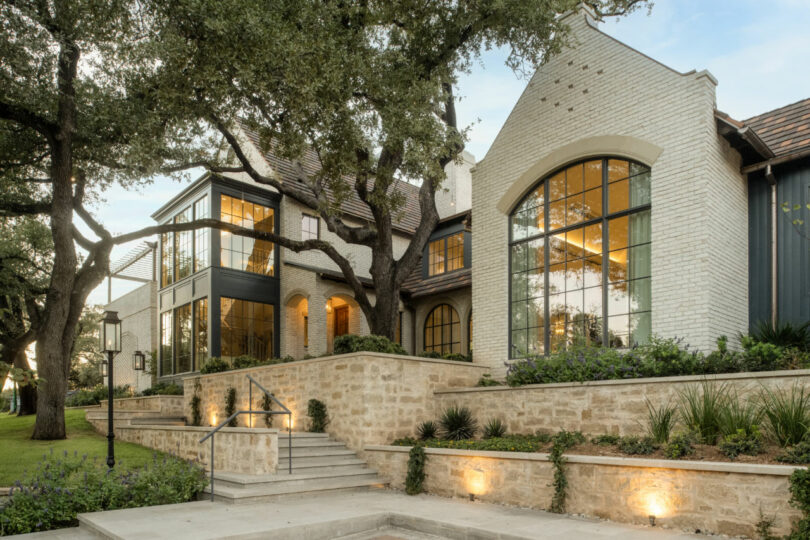 Two-story modern house with large windows, light brick exterior, stone retaining wall, and landscaped yard with mature trees—perfect for your Friday 5 gatherings.