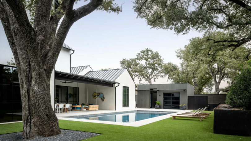Modern backyard with a rectangular swimming pool, lounge chairs, and a covered patio area next to a white house surrounded by large trees—perfect for relaxing or hosting a Friday 5 gathering with friends.