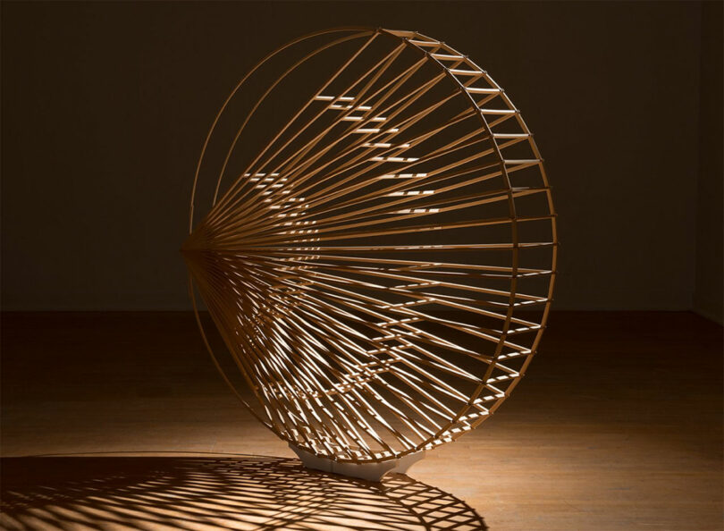 A large wooden geometric sculpture by Vivian Chiu, with radiating spokes, stands on a wooden floor, casting intricate shadows in a dimly lit room.