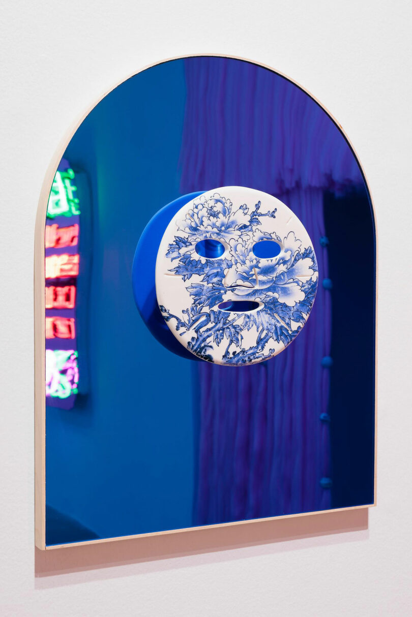 A blue, arched mirror by Vivian Chiu features a white porcelain mask with floral designs at its center. Neon lights and draped fabric are reflected in the mirror.