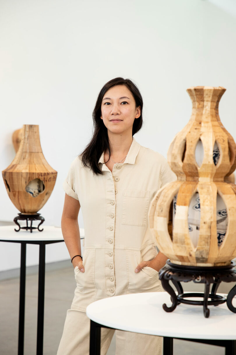 A woman in a light-colored jumpsuit stands with hands in pockets between two wooden sculptural vases by Vivian Chiu, displayed on round white tables in a gallery setting.