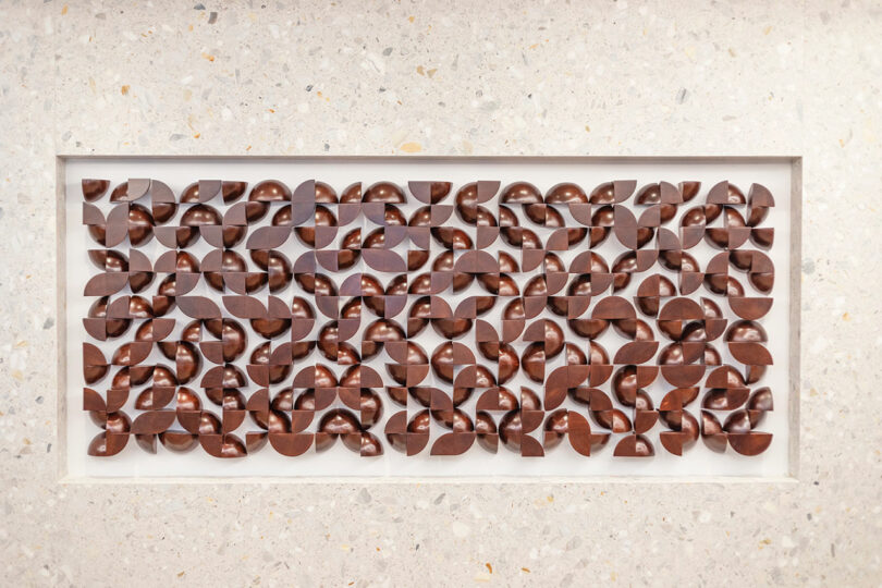 Rectangular wooden wall art by Vivian Chiu features repeating geometric, leaf-like shapes arranged in a grid pattern, embedded in a light-colored textured surface.