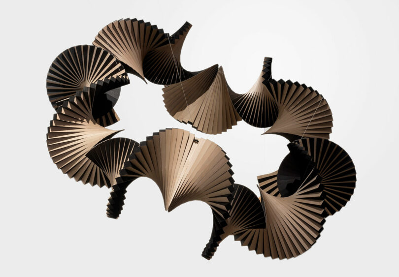A sculptural light fixture by Vivian Chiu, featuring interlocking, accordion-like pleated forms arranged in a circular pattern, suspended against a plain white background.