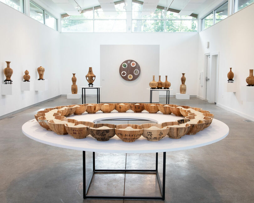 A contemporary gallery displays wooden bowls arranged in a circle on a table, with vases on pedestals and a round Vivian Chiu artwork on the wall in the background.