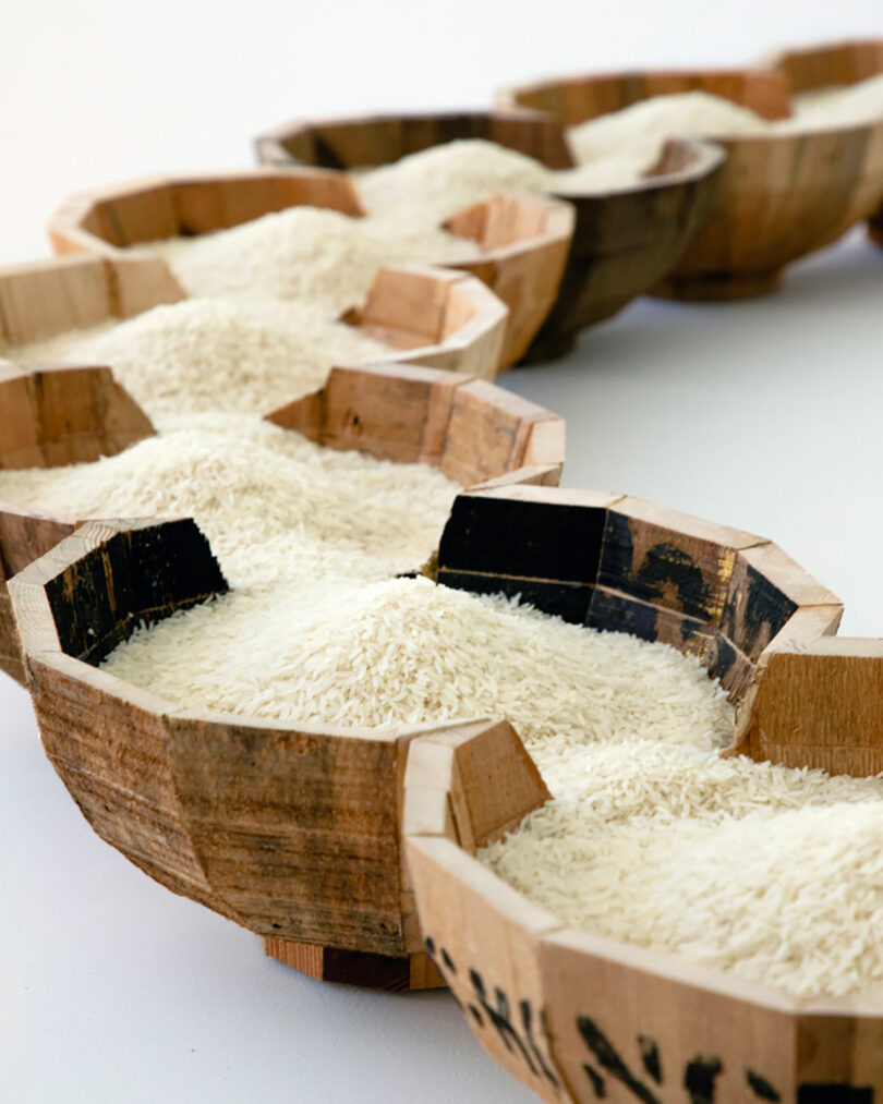 Several wooden bowls filled with uncooked white rice, reminiscent of Vivian Chiu’s intricate arrangements, are displayed in a zigzag pattern on a white surface.