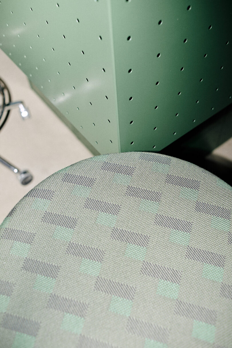 A close-up of a patterned green chair seat by Form Us With Love sits next to a light green perforated metal panel, with part of a chrome chair leg visible in the background.