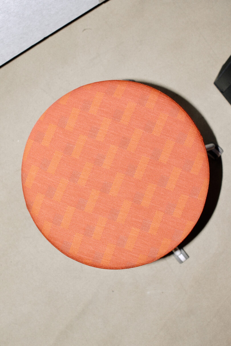 Overhead view of a round stool seat upholstered in orange houndstooth fabric, with chrome hardware visible on the side—a contemporary design by Form Us With Love.
