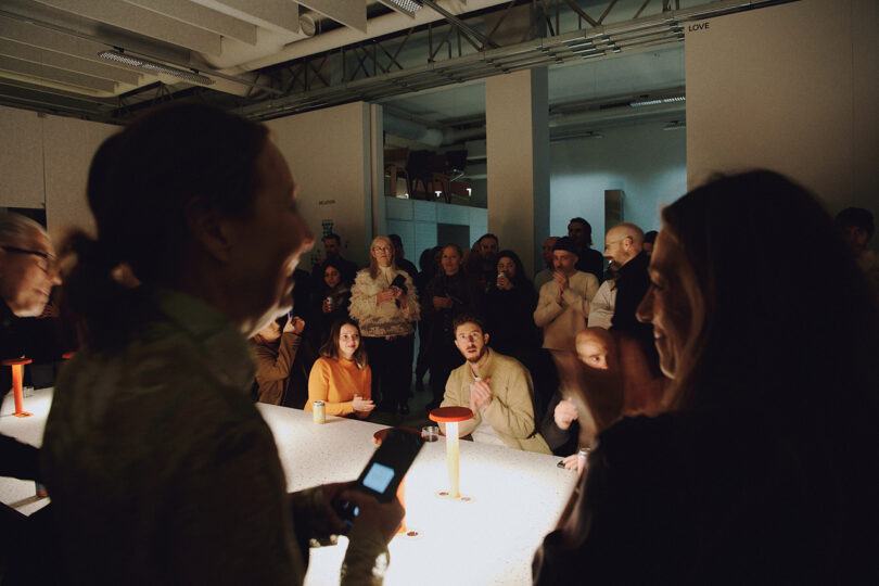 A group of people are gathered around a lit Form Us With Love table in a dimly lit room, some sitting and some standing, engaging in conversation and observing.