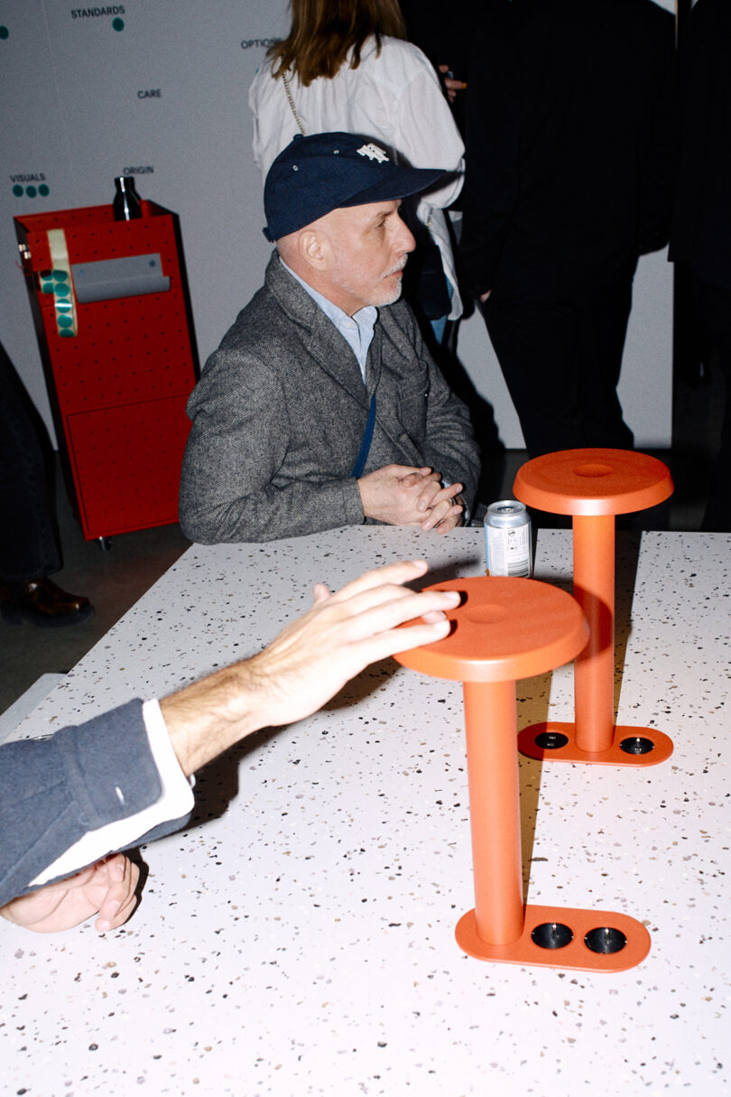 A man in a gray coat and blue cap sits at a speckled table with orange Form Us With Love stands, while another person's hand reaches across the table.