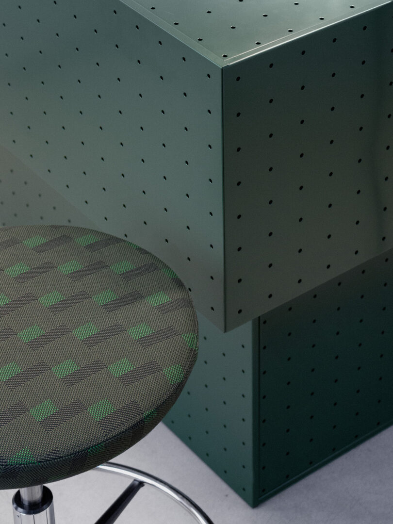 A close-up of a green patterned stool designed by Form Us With Love sits next to a green, perforated metal box, both on a light gray surface.