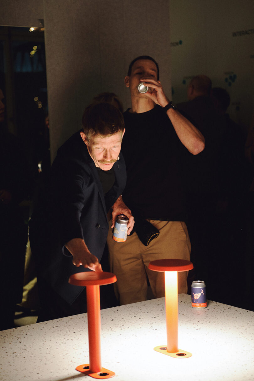 Two people stand at a table with illuminated orange poles by Form Us With Love, one reaching towards a pole and the other drinking from a can. Several other people are visible in the background.