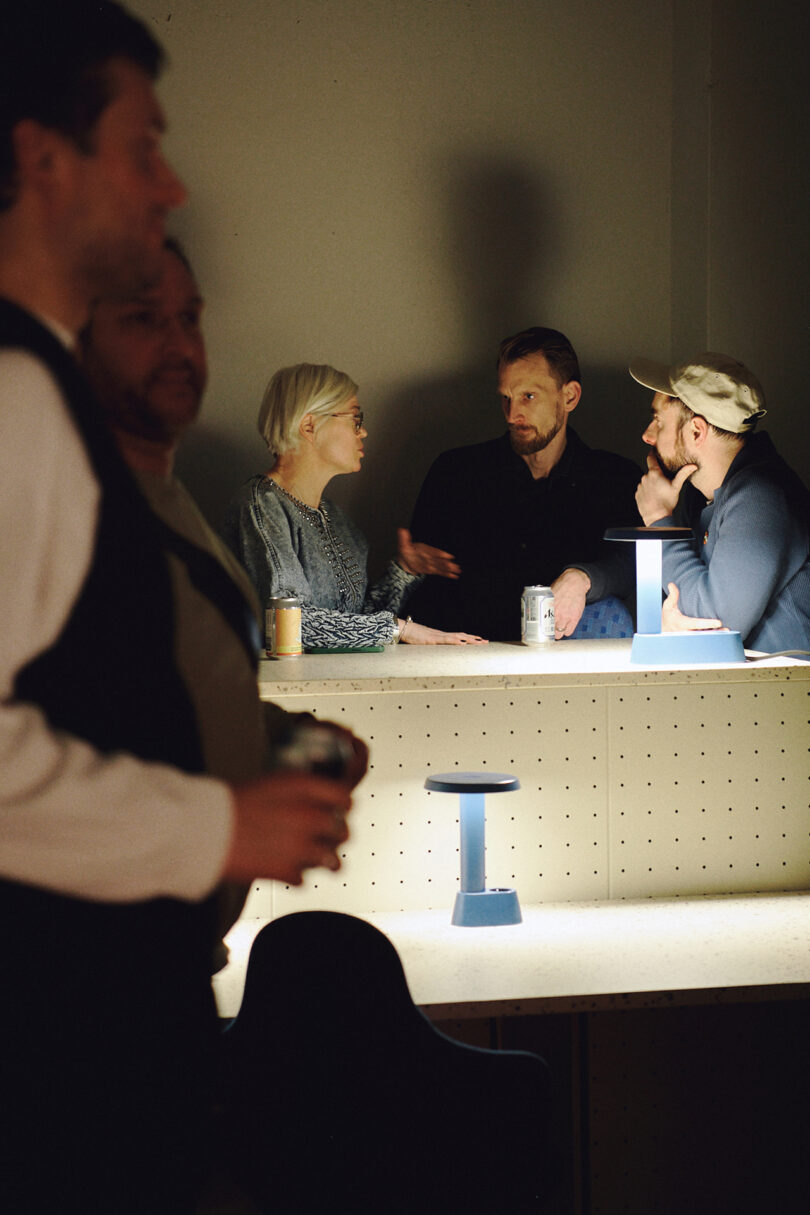 Four people sit and talk at a small table with blue lamps designed by Form Us With Love, while two others stand in the foreground, partially out of focus.