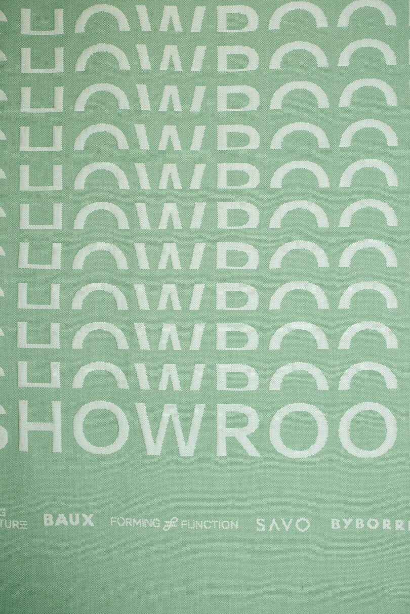 Green fabric or banner with a repeating white "SHOWROOM" text pattern, featuring brand logos such as BAUX, Form Us With Love, Forming & Function, SAVO, and BYBORRE along the bottom.