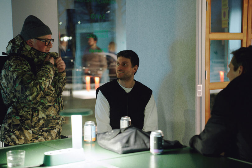 Three people are gathered around a green table indoors, talking. There are cans, a backpack, and a jacket on the table—creating an atmosphere reminiscent of a creative session at Form Us With Love. One person stands while two are seated.