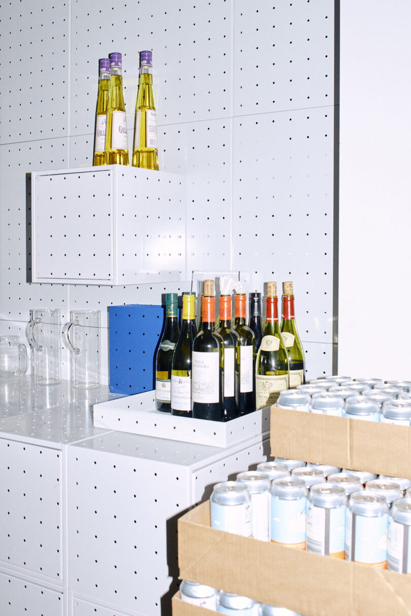 Shelves by Form Us With Love hold bottles of wine and olive oil, clear pitchers, a blue box, and stacked boxes of canned drinks in a bright, organized space with white pegboard walls.