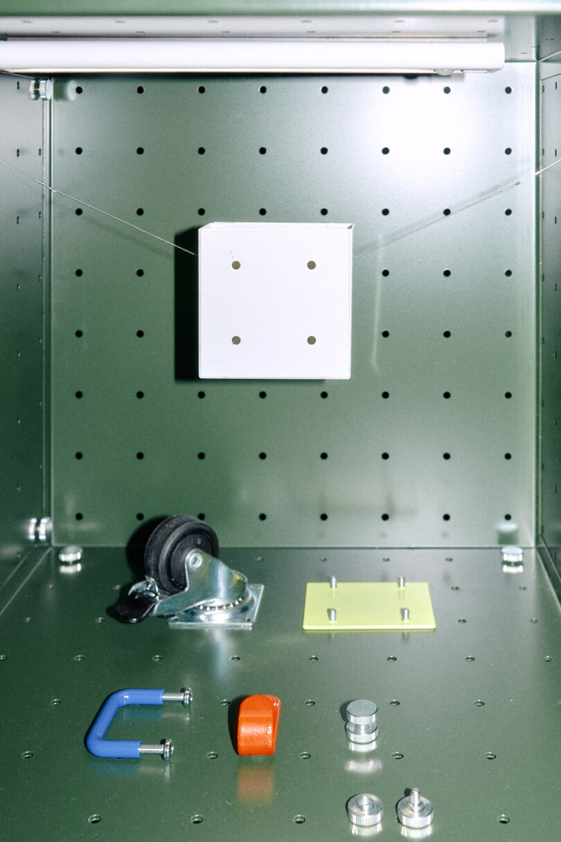 Interior of a green metal storage cabinet with a perforated back panel, fluorescent light, and various hardware components including a caster wheel, clamp, and fasteners—a practical design inspired by Form Us With Love.