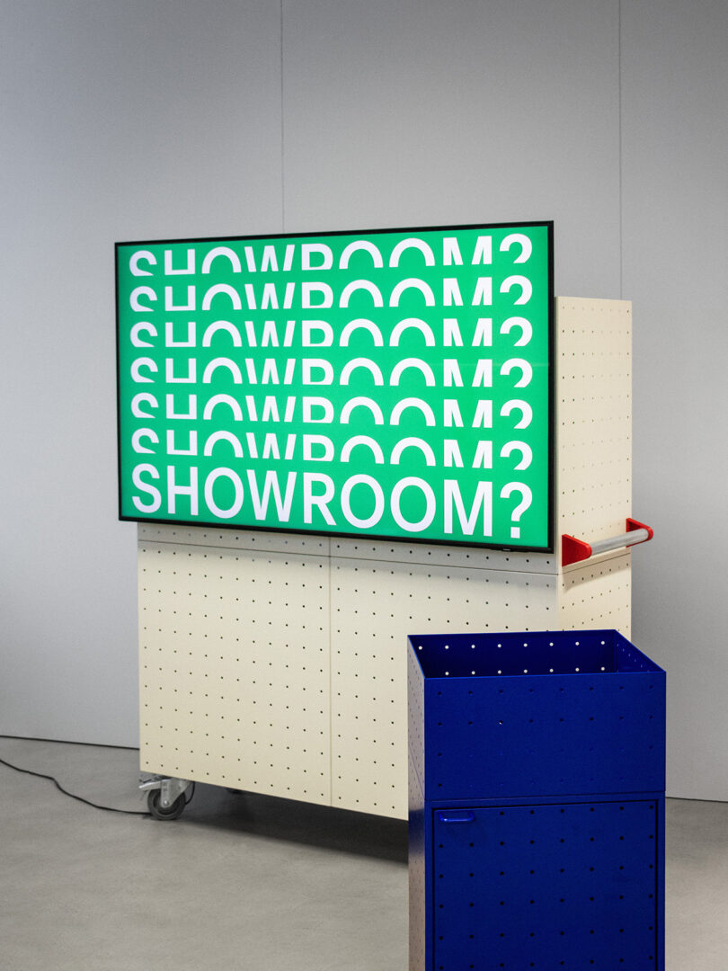 A digital screen by Form Us With Love displays the word “SHOWROOM?” in white text on a green background, mounted on a pegboard cart beside a blue box on a gray floor.