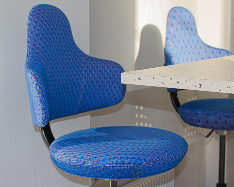 Two blue upholstered chairs with curved backs by Form Us With Love are positioned next to a white speckled tabletop, with sunlight casting shadows on the wall.