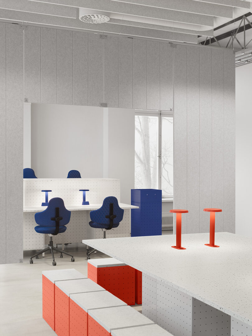 Modern office space with grey walls, large window, blue office chairs, white desks, and red stools; geometric furniture and minimal decor inspired by Form Us With Love.