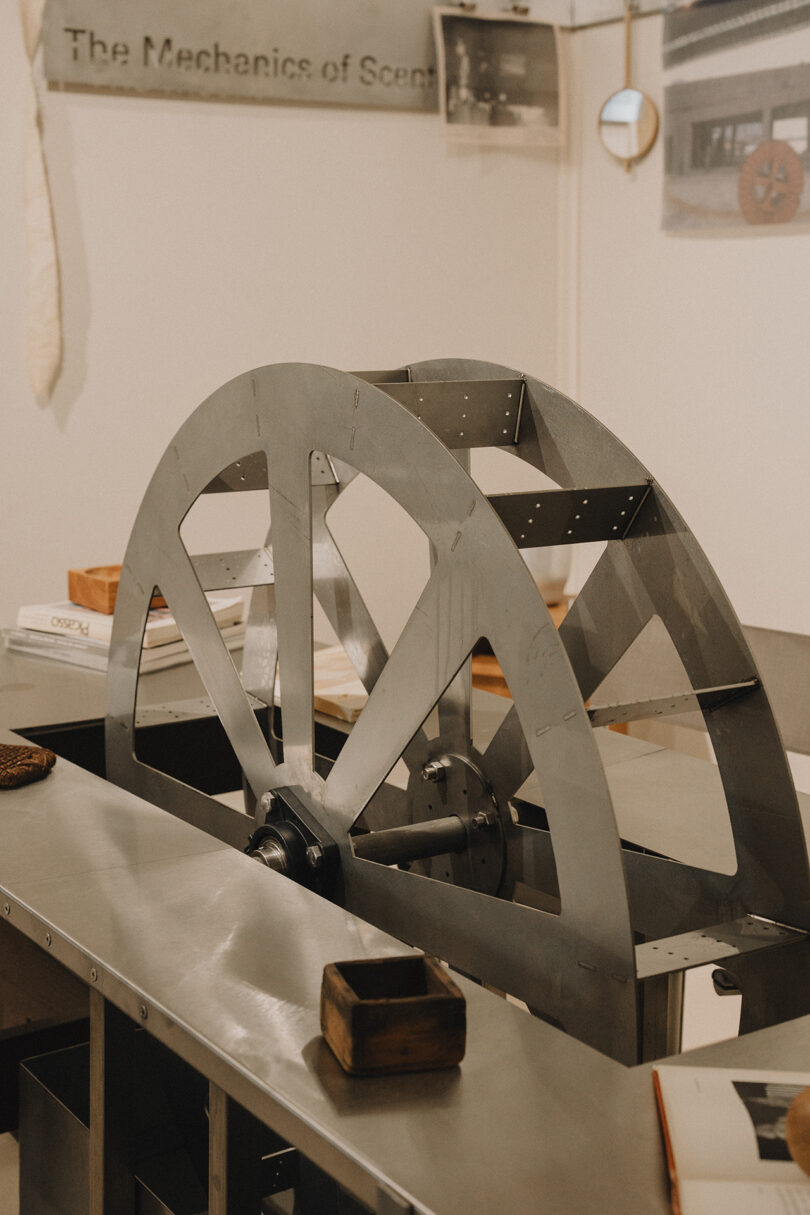 A large metal wheel is mounted on a table surrounded by books and objects in an exhibition space with a sign reading "The Mechanics of Scene.