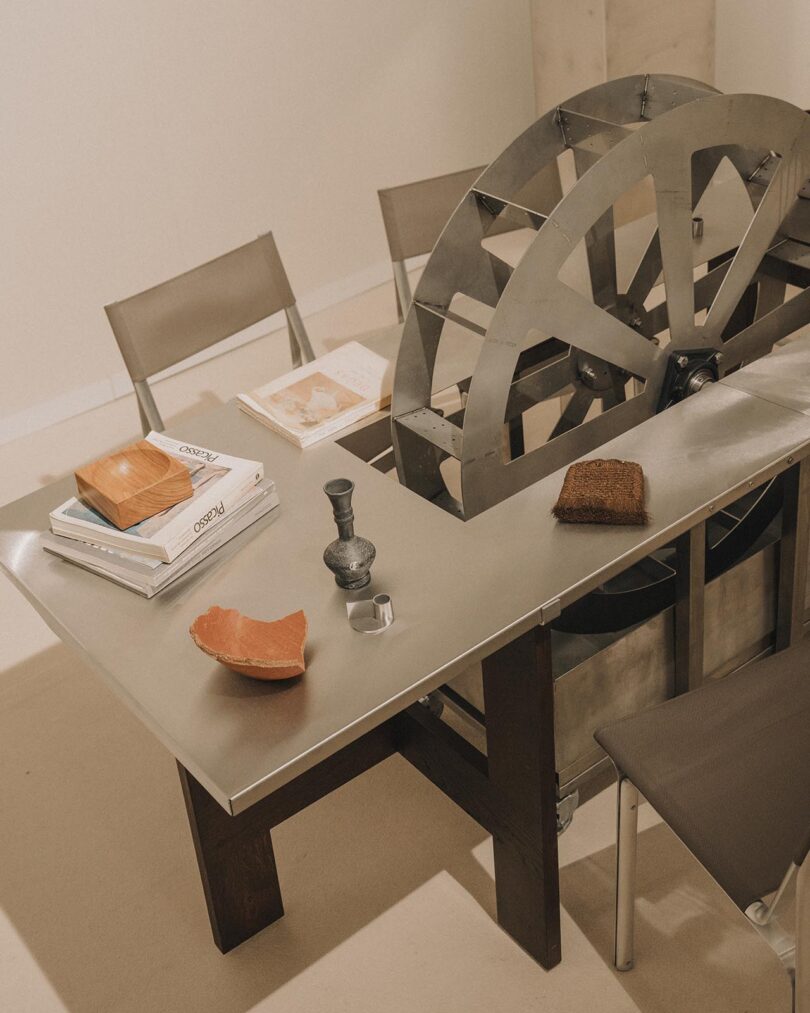 A metal table with books, a mortar and pestle, bread, a cup, pottery fragments, and a large metal wheel centerpiece, surrounded by four chairs in a minimalist setting.