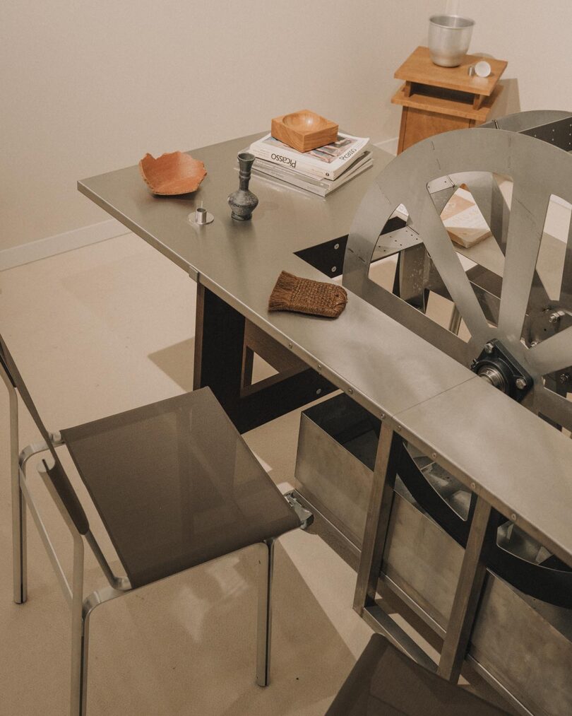 A modern room with a glass chair, a metal table featuring an industrial fan component, books, a bowl, and decorative objects on various surfaces.