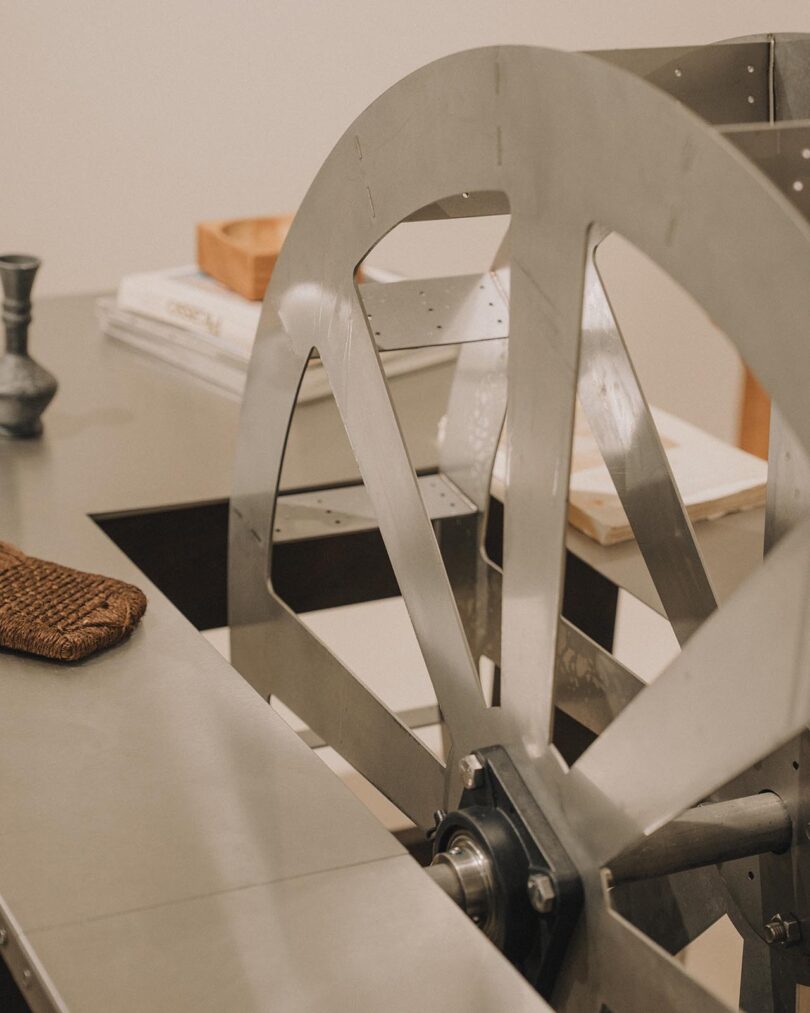 A large metal wheel is attached to a piece of equipment on a table, with a woven glove, candle holder, and stacked books in the background.
