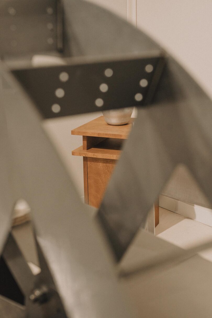 A close-up of a metallic structure with circular holes partially obscures a wooden side table and a ceramic bowl in the background.