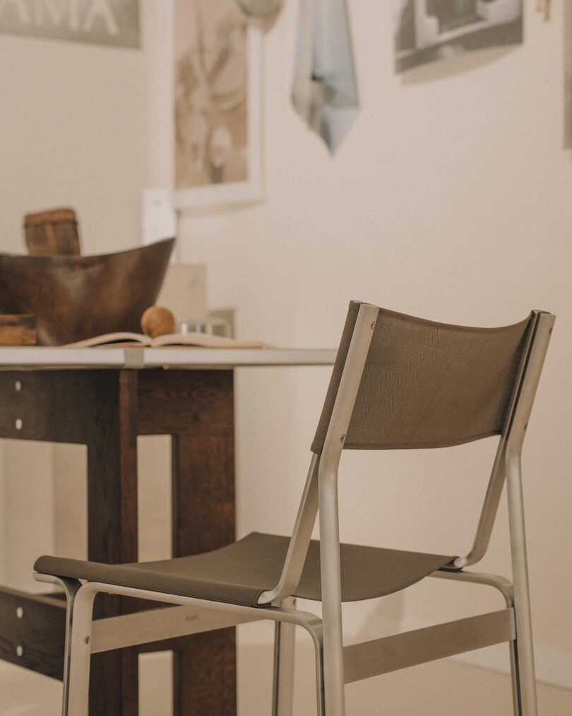 A metal and fabric chair is positioned next to a wooden desk with a bowl and scattered items; framed pictures hang on the wall in the background.