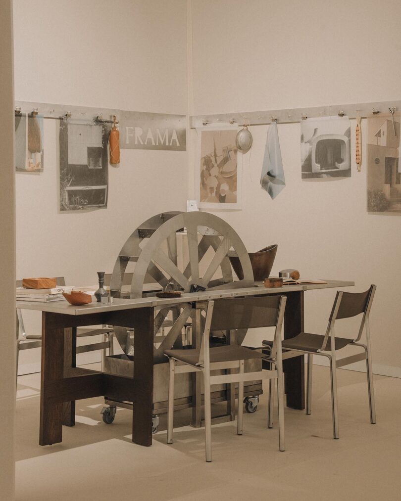 A minimalist room features a metal table with a large wheel at its center, surrounded by chairs; artwork, objects, and textiles hang on the beige walls, with a "FRAMA" sign visible.