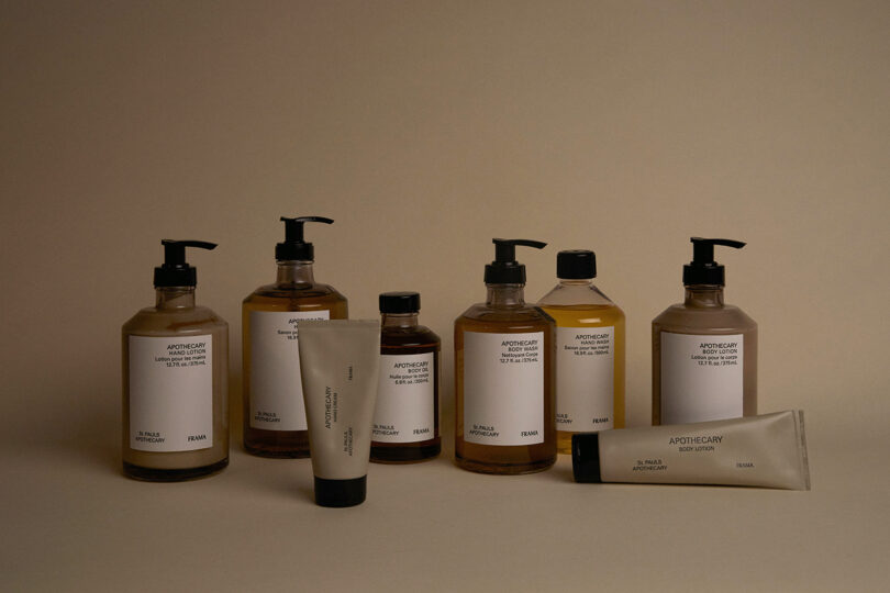 Seven bottles and two tubes of apothecary-style personal care products with minimalist white labels are arranged on a beige surface against a beige background.