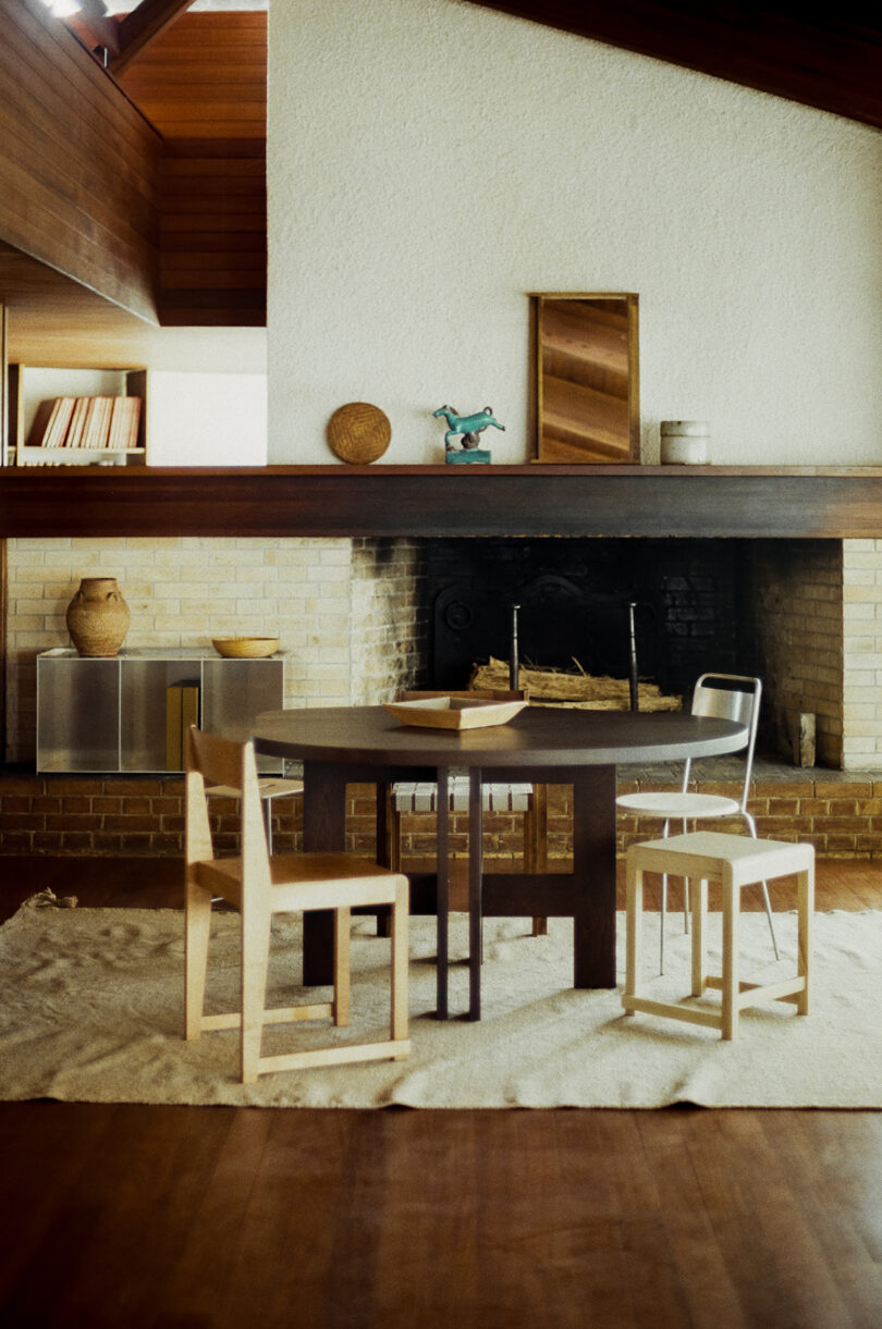 A round wooden table with four chairs sits on a rug in front of a brick fireplace in a modern living room with wooden floors and shelves.