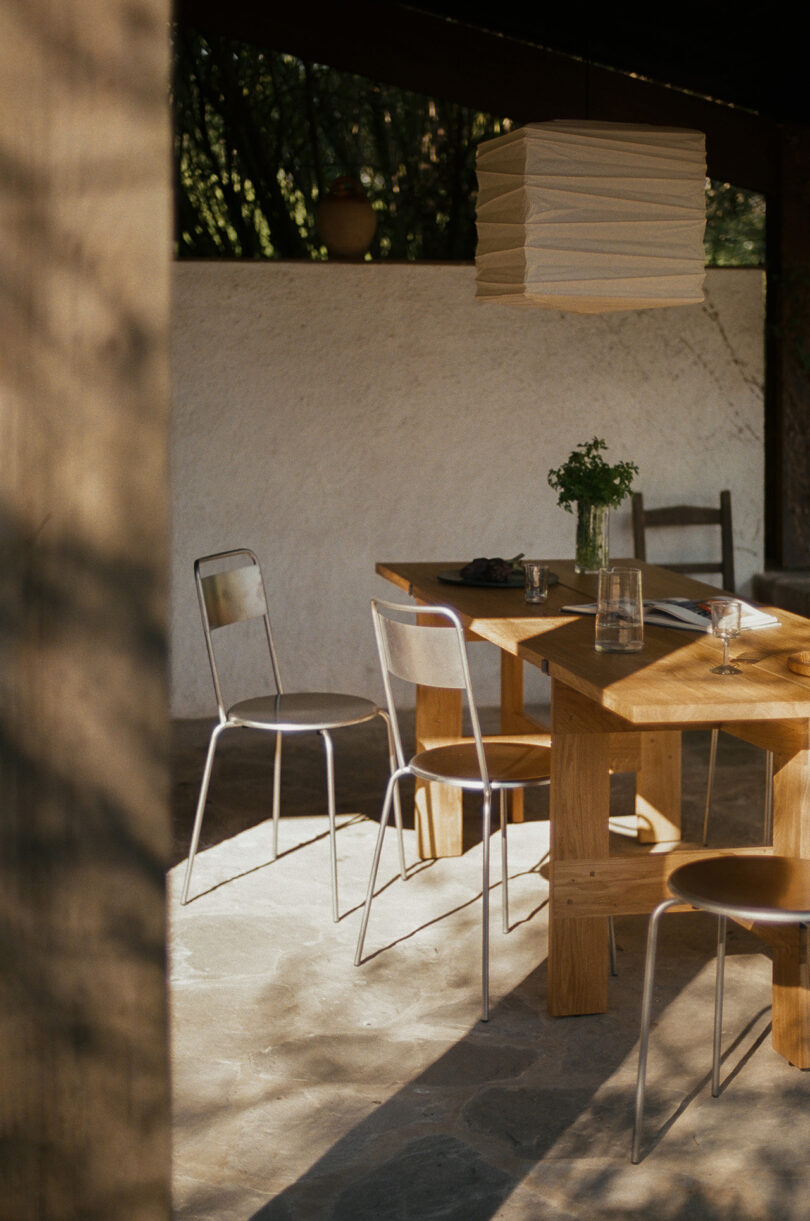 A wooden dining table with clear chairs and a paper lantern pendant lamp, set on a stone floor in a sunlit, partially outdoor space.