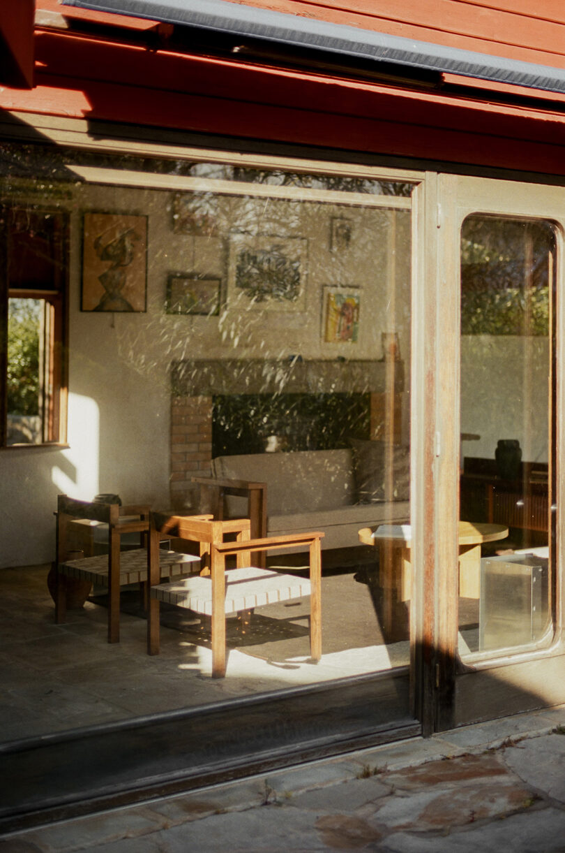 A sunlit room with wooden chairs and a table is visible through large glass doors; framed artworks hang on the white walls inside.