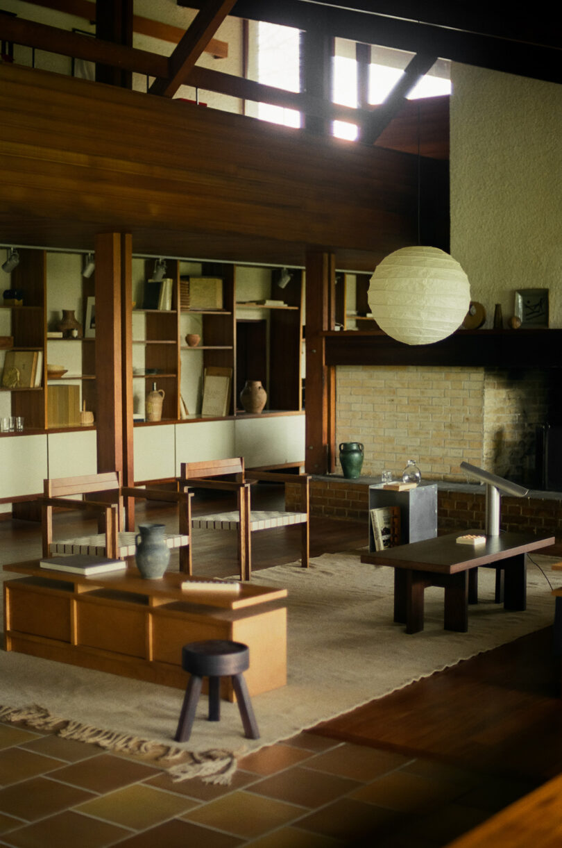 A mid-century modern living room with wooden furniture, a brick fireplace, built-in shelves, and a large round paper lantern hanging from the ceiling.