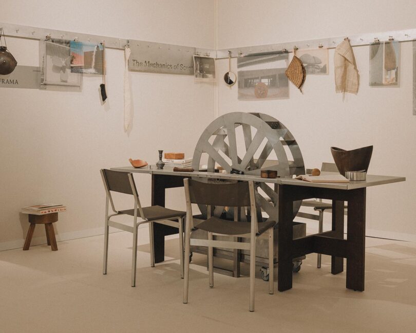 A metal table with assorted dishes and utensils is surrounded by metal chairs; a large metal wheel is mounted at one end. Various objects and photos are displayed on the wall behind.