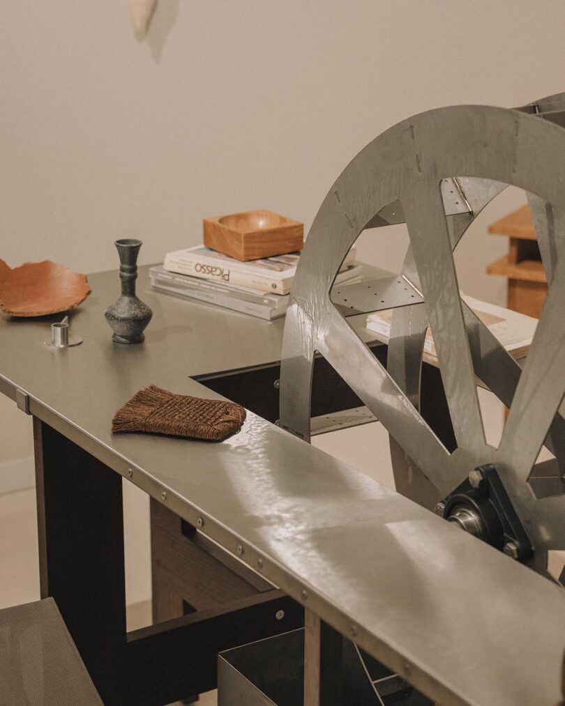 A large metal wheel is mounted on a table beside a small stack of books, a wooden tray, a metal vase, and a piece of brown fabric in a minimalist room.