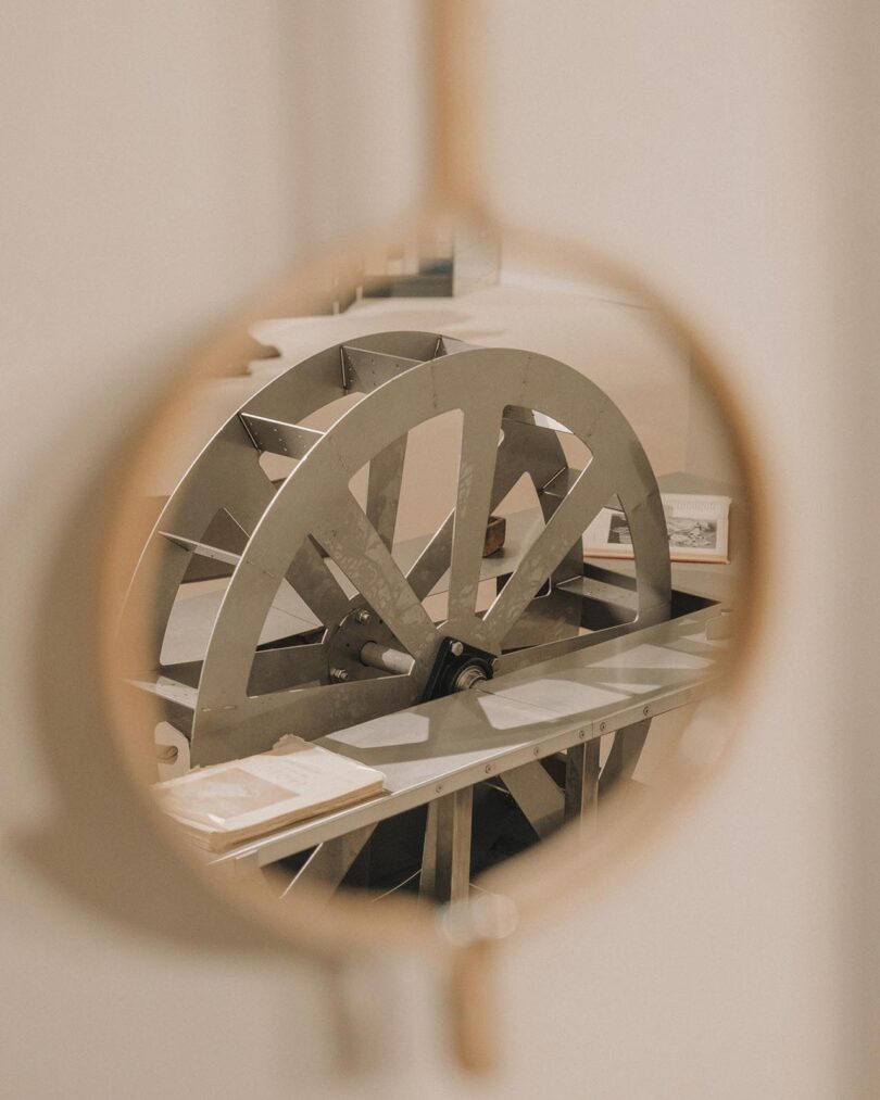 A large metal gear or wheel is seen through an oval-shaped mirror, with part of a table and an open book visible in the background.