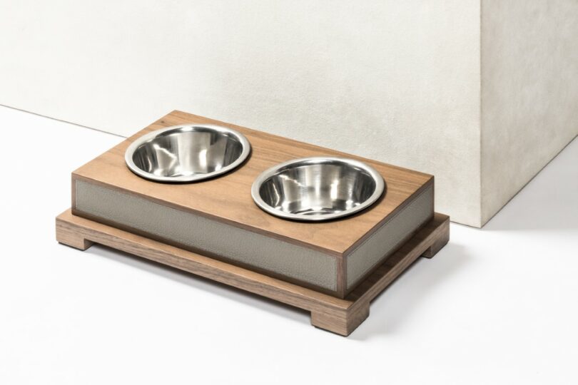 A wooden pet feeder with two stainless steel bowls is placed on a white surface near a light-colored wall