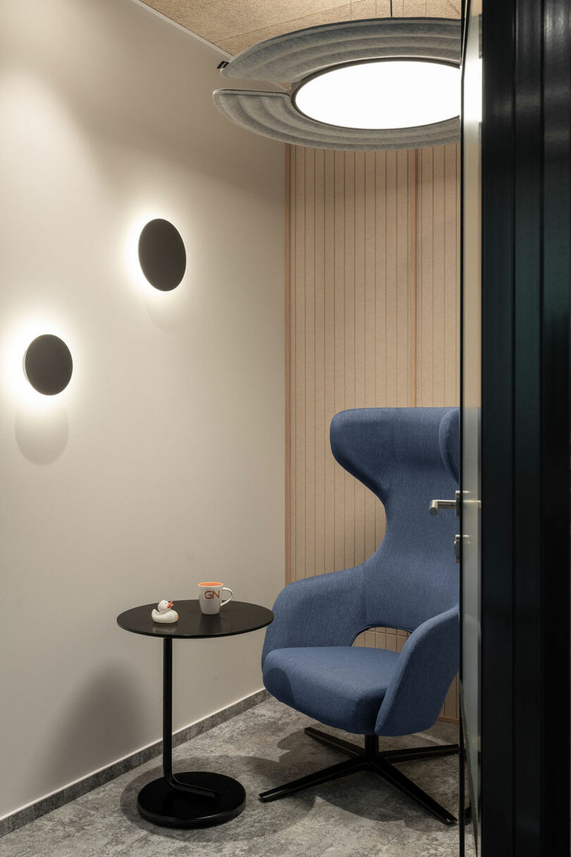 A modern BIT CREATIVE office nook with a blue lounge chair, a round black side table holding a mug and small plate, circular wall lights, and a ceiling light.