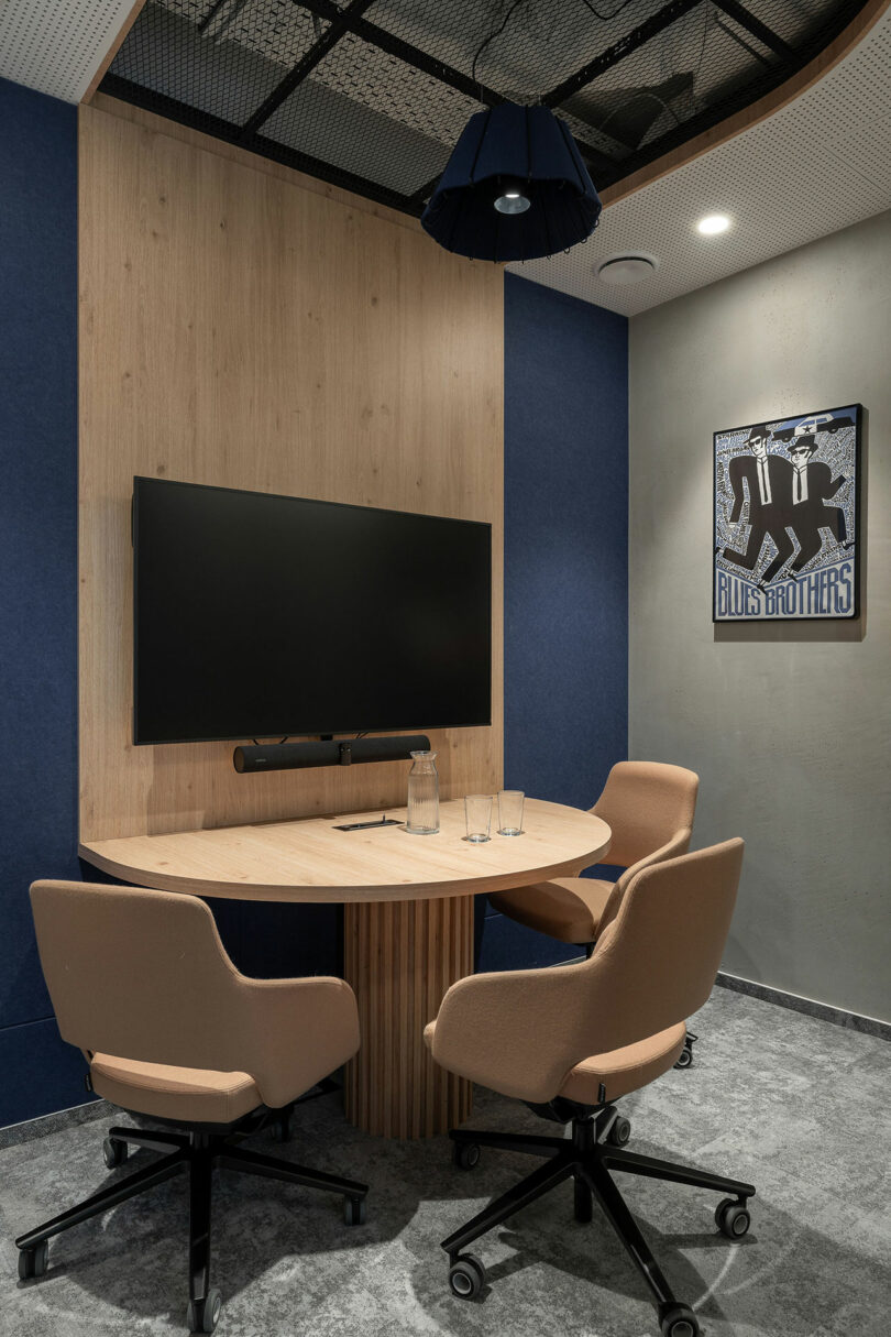 A small meeting room by BIT CREATIVE with a round wooden table, three beige chairs, a wall-mounted TV, a water carafe with glasses, and a "Blues Brothers" poster on the wall.