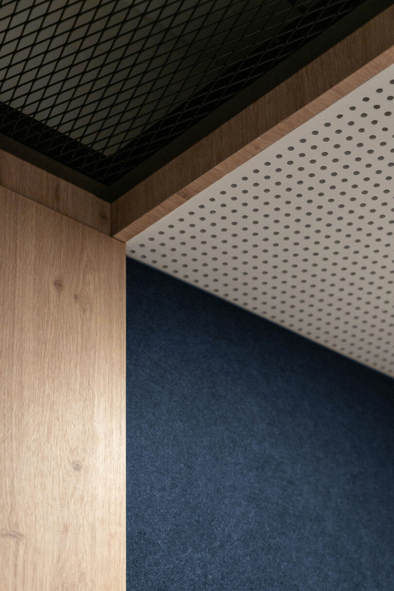 A close-up of an interior corner by BIT CREATIVE, featuring a wooden panel, a perforated white panel, a blue textured wall, and a black mesh metal grid overhead.