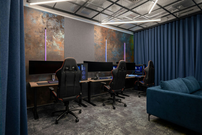 A modern gaming room by BIT CREATIVE features three ergonomic chairs at desks, large monitors and PCs, a blue sofa, blue curtains, and contemporary lighting.