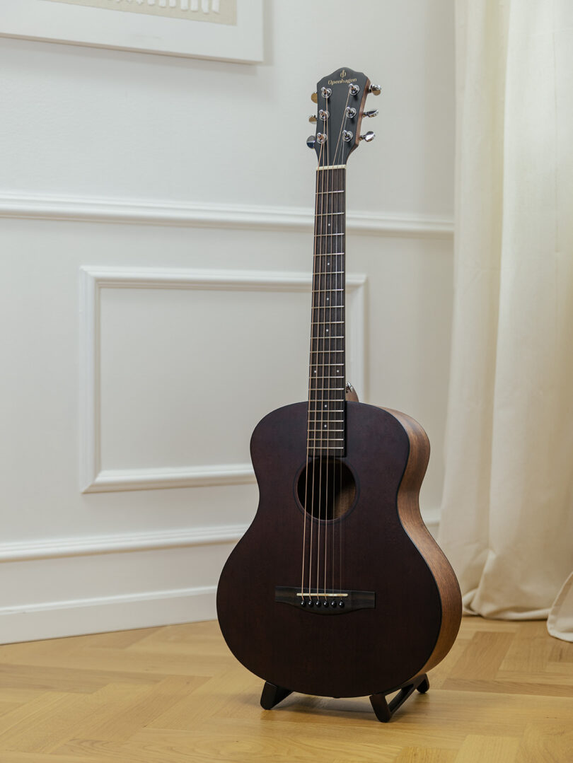 A dark brown acoustic guitar with six strings is standing on a small stand on a wooden floor in a room with white walls and decorative molding.