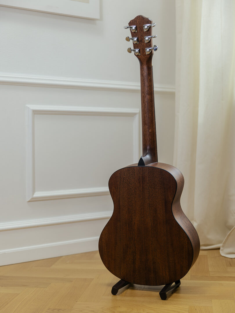 A wooden acoustic guitar shown from the back, standing on a black stand on a wooden floor near a cream-colored wall and curtain.