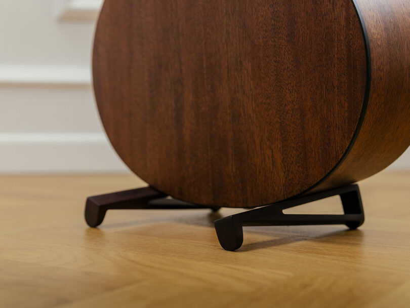 A close-up of a round wooden speaker or electronic device on a stand, placed on a hardwood floor near a white wall.