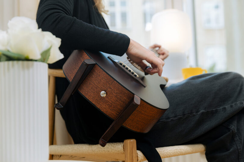 A person sitting on a chair strums an acoustic guitar indoors, next to a white vase of flowers and a yellow mug.