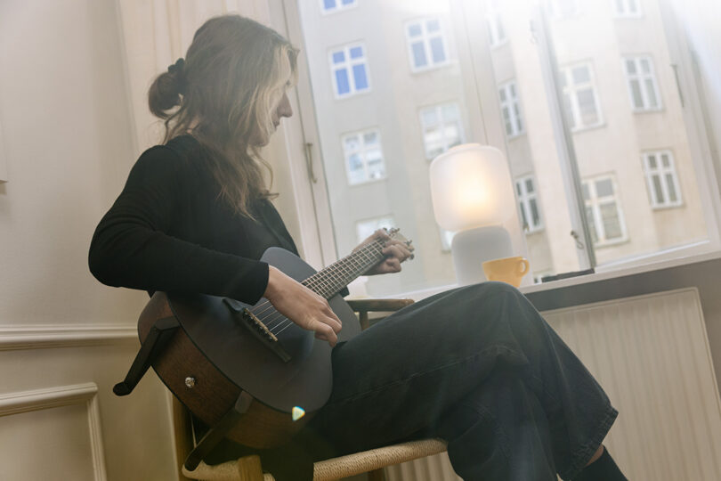 Person sitting on a chair indoors playing an acoustic guitar near a window with sunlight shining in and a cup and lamp on the windowsill.